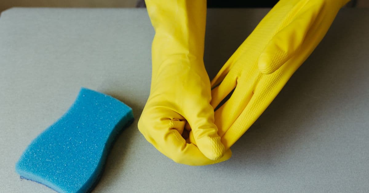 Mãos com luvas de borracha amarelas ao lado de uma esponja azul sobre uma bancada cinza
